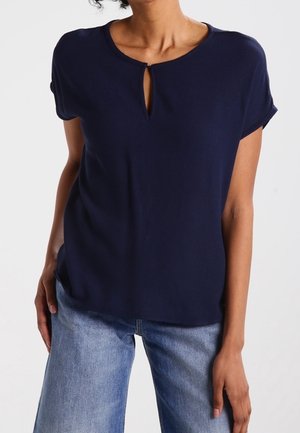 Top blu navy a maniche corte realizzato in tessuto morbido, con dettaglio a chiave sul collo e una vestibilità rilassata. Abbinato a jeans chiari.