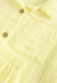 Gul button-up skjorte med struktureret stof, brystlomme og rynket taljedetalje. Træknapper fremhæver designet.