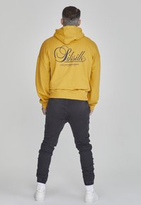 Senfgelbes Hoodie mit einem großen schwarzen Logo und Text auf dem Rücken, kombiniert mit schwarzen tapered Pants und weißen Sneakern. Schlichter Hintergrund.