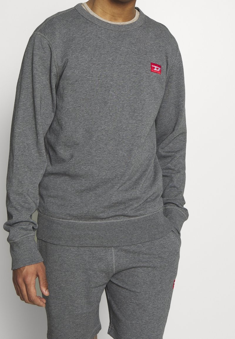 Homme portant un sweat-shirt gris à manches longues et un short assorti avec un petit logo rectangulaire rouge sur la poitrine et le short, les mains dans les poches.