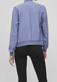 Ljuslila bomberjacka med ribbad krage och mudd, med en mjuk textur och elastisk midja. Bärs över svarta byxor.