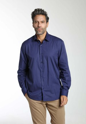 Homme aux cheveux courts et foncés avec une barbe, portant une chemise bleu marine boutonnée et un pantalon beige, debout avec une main dans la poche.