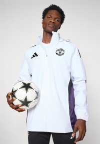 White athletic jacket with black accents and Manchester United logo, featuring a hood and elastic cuffs. Model holds a patterned soccer ball.