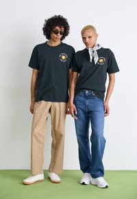 Sorte grafiske t-skjorter med sol-design, beige bukser og blå jeans. Skoene inkluderer hvite joggesko og platåskinn. Lys bakgrunn.