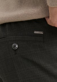 Pantaloni a scacchi scuri con chiusura a bottone, tasca frontale e etichetta metallica con la scritta "JACK & JONES," abbinati a un maglione beige.