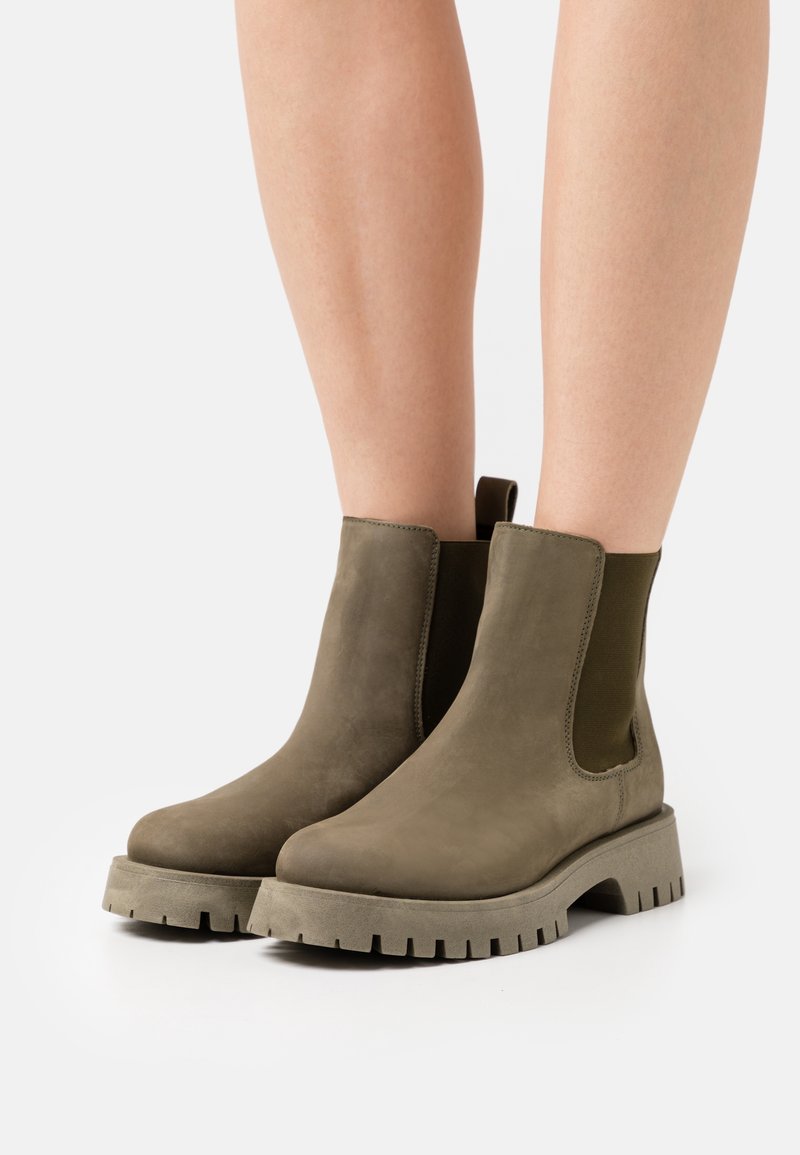 Bottines à hauteur de cheville en daim vert olive avec semelles épaisses portées sur des jambes nues sur un fond uni.
