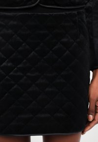 Jupe noire matelassée avec couture en losange portée par une personne, montrant une main reposant à côté de l'ourlet de la jupe.