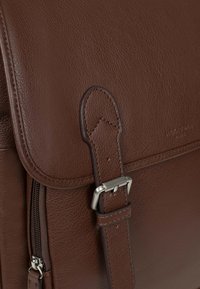 Brązowa skórzana torba o gładkiej fakturze, z klapką zamykaną na zatrzask oraz srebrnymi metalowymi dodatkami na regulowanym pasku. Widoczny detal w postaci zamka błyskawicznego.