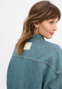 Veste en denim bleu moyen avec une coupe décontractée. Dispose d'une étiquette à l'arrière et de coutures visibles, mettant en avant un design classique.