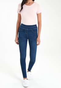 Jeggings bleu taille haute avec des détails de couture, deux poches avant, associés à une chemise à manches courtes rose clair et des baskets blanches.