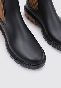 Bottes en caoutchouc noires à chevilles avec des panneaux élastiques en tan striés, dotées d'une semelle épaisse et texturée et d'un design à bout rond.