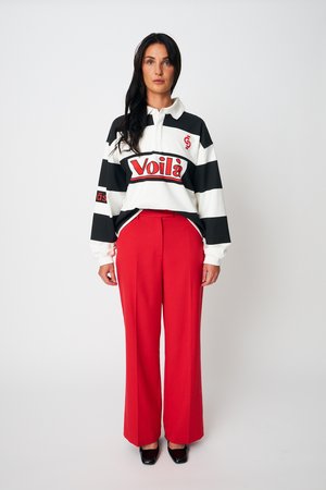 Pikajuustega naine, kes kannab must-valget triibulist pika varrukaga särki, millel on "Voilà" logo, ning erkpunaseid laia säärega pükse, seisab sirgelt.