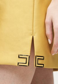 Κοντινό πλάνο στο κάτω μέρος κίτρινης μουσταρδί φούστας με κεντρική σχισμή, περιτριγυρισμένη από δύο χρυσές και μαύρες γεωμετρικές μεταλλικές λεπτομέρειες, δίπλα σε ένα χαλαρό χέρι.