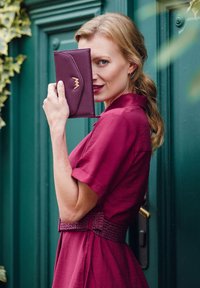 Burgundy wallet with curved flap and golden accent held against a model's face. She wears a matching dress with textured belt against a green door.