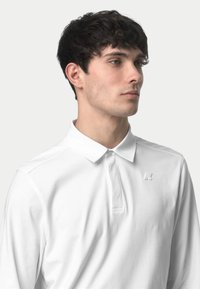 Polo shirt bianco a maniche lunghe con colletto, dotato di una placca con mezzi bottoni e un sottile dettaglio del logo sul petto. Tessuto morbido e liscio.