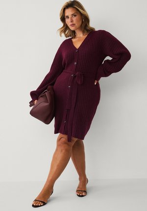 Woman wearing a burgundy ribbed knit button-up dress with a belt, holding a matching bag and wearing black open-toe heels, standing against a plain wall.