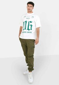 Balts T-krekls ar zaļu grafisko dizainu, kurā redzami "06" un "SIXTH JUVE". Olīvu zaļas cargo bikses ar sānu kabatām. Baltas tenisītis.