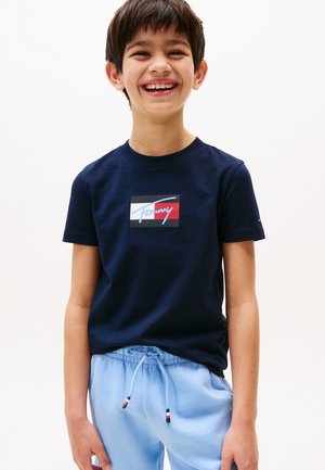 Ragazzo sorridente che indossa una t-shirt blu navy con un logo rosso, bianco e nero e pantaloni della tuta azzurri con coulisse, su sfondo bianco.