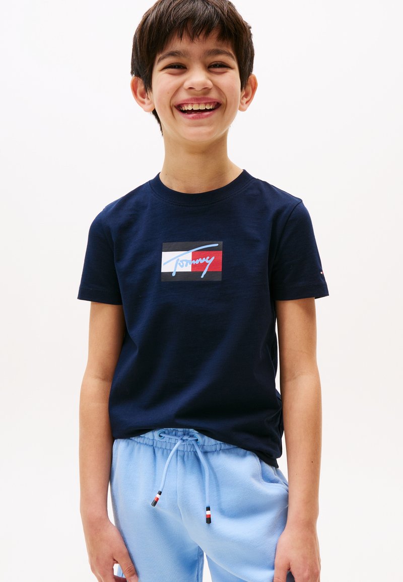 Ragazzo sorridente che indossa una t-shirt blu navy con un logo rosso, bianco e nero e pantaloni della tuta azzurri con coulisse, su sfondo bianco.