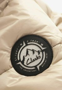 Patch rond noir et gris avec le logo Clarks et un motif montagne sur tissu matelassé beige, comportant des coordonnées et le texte « L’aventure commence ici ».