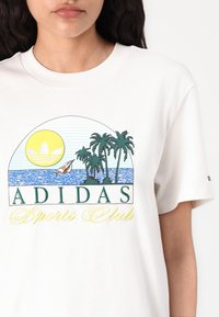 White cotton t-shirt featuring a colorful graphic of a sun, palm trees, and a sailboat, with "ADIDAS Sports Club" text in green and yellow.