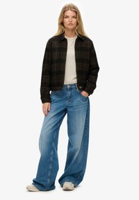 Zwarte en bruine geruite jas over een lichtgrijs topje, gecombineerd met brede blauwe jeans en beige sneakers. Rechte snit en ontspannen pasvorm.