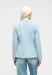 Γυναίκα με μακριά ξανθά μαλλιά, φοράει ανοιχτό μπλε σακάκι και ξεθωριασμένο μπλε τζιν, γυρισμένη πλάτη σε έναν απλό λευκό φόντο.