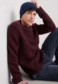 Burgundijas adīta džemperis ar ribotām piedurknēm un apakšmalu, apvienots ar teksturētiem tumši ziliem kordūra biksēm. Modelis valkā tumši zilu ribotu adītu cepuri.