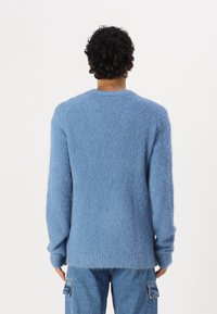 Αχνό, ανοιχτό μπλε πουλόβερ με μακριά μανίκια και στρογγυλή λαιμόκοψη. Διαθέτει χαλαρή εφαρμογή και υφή. Φορεμένο πάνω από μπλε τζιν παντελόνι.