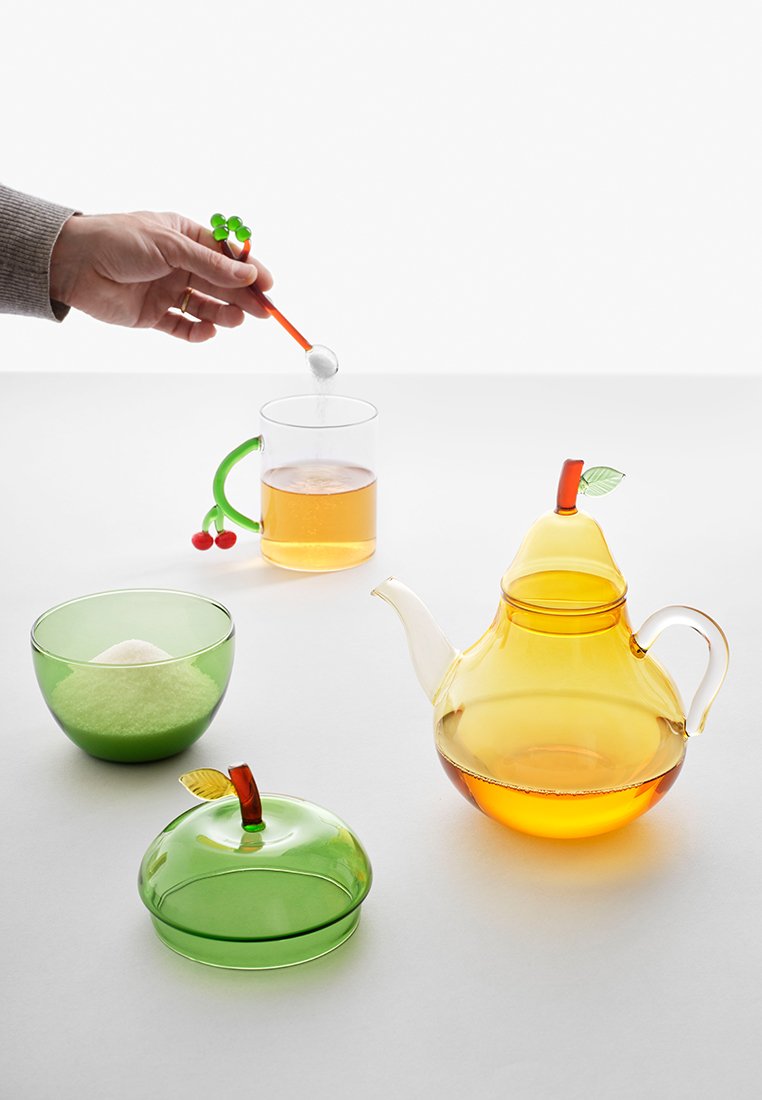 Glazen theepot in het geel, met een bladaccent; groene kom met suiker; doorzichtige mok met thee; kleurrijke lepel met een kersontwerp.