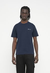 Μπλε ναυτικού βαμβακερό t-shirt με στρογγυλή λαιμόκοψη, κοντά μανίκια και μικρό λευκό λογότυπο στο στήθος. Φορώντας το με μαύρο παντελόνι.