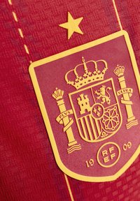 Un tissu rouge avec un motif texturé arbore un blason brodé jaune, une étoile et des accents décoratifs, mettant en valeur l'emblème de l'Espagne.