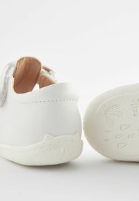 Białe, miękkie buty z imitacji skóry z teksturowaną gumową podeszwą. Zauważalne szczegóły w kształcie kocich uszu na pięcie oraz minimalistyczny design.