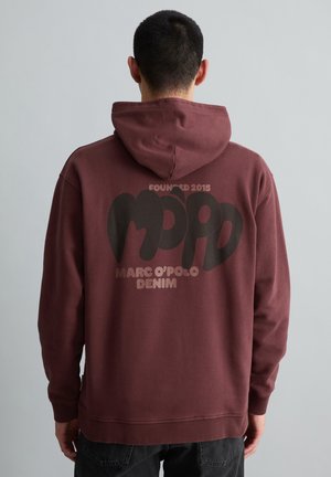 Mann trägt einen weinroten Hoodie mit dem Schriftzug "Founded 2015 Marc O'Polo Denim" und einer Grafik auf dem Rücken, steht vor einem einfarbigen grauen Hintergrund.