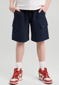 Navy cotton cargo shorts with two side pockets, elastic waistband, and visible stitching. Paired with red and cream high-top sneakers.