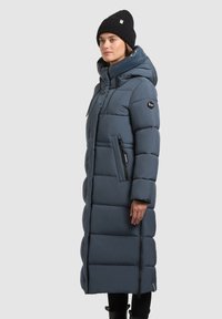 Ilgas, pūkuotas, tamsiai mėlynas žieminis paltas su kapišonu, užtrauktukais kišenėse, siuvinėtu dizainu ir juodu megztu kepurėle su raukšlėmis.