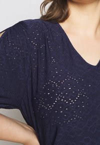 Námořnická modrá krajková blůza s výstřihem do V, zdobená vyřezávanými květinovými vzory a krátkými rukávy. Měkká textura s mírnou průsvitností.