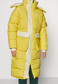 Keltainen toppatakki valkoisilla yksityiskohdilla ja tikattu etuosa. Varustettu isoilla taskuilla, korkealla kaulukalla ja vetoketjulla. Kuvioitu kangas.