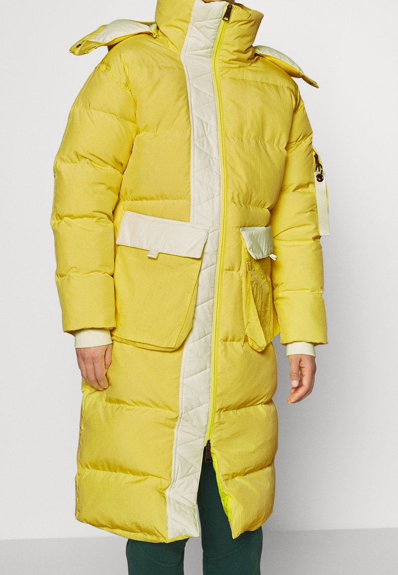 Keltainen toppatakki valkoisilla yksityiskohdilla ja tikattu etuosa. Varustettu isoilla taskuilla, korkealla kaulukalla ja vetoketjulla. Kuvioitu kangas.