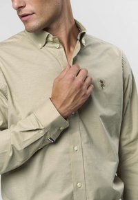 Beiges Hemd zum Knöpfen aus glattem Stoff, mit einem kleinen gestickten Logo auf der Brust und einem dezenten Streifenmuster an den Manschetten.