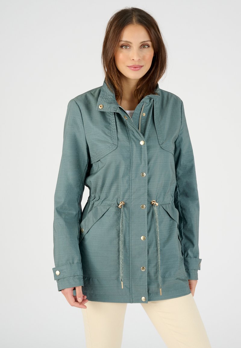 Veste légère en bleu sarcelle avec fermeture éclair et pression, taille cintrée, grandes poches avant et capuche, présentant un motif texturé subtil.