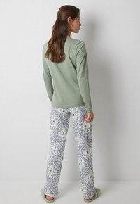 Langärmliges, weiches grünes Oberteil kombiniert mit leichten, gemusterten Hosen mit blauen Akzenten und floralen Designs; Modell steht mit dem Rücken zur Kamera.