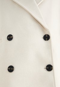 Manteau croisé blanc avec quatre boutons noirs et un large col à revers cranté.