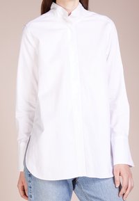 Mujer vestida con una camisa blanca de manga larga con cuello alzado y jeans de mezclilla azul claro, de pie contra un fondo beige.