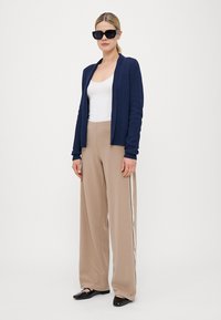 Marineblå strikket cardigan over hvit tank top, parret med beige vide bukser med side-striper, og sorte spisse ballerinas. Motebeskyttelsesbriller på.
