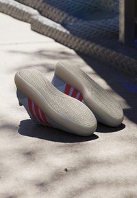 Sportske cipele u sivoj boji s crvenim akcentima. Debeli, teksturirani gumeni đonovi. Mekani gornji materijal. Glatka završna obrada s tri dijagonalne pruge.