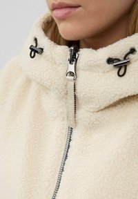 Nahaufnahme einer Person, die eine cremefarbene Sherpa-Jacke mit einem silbernen Reißverschluss und schwarzen Kordelzug-Toggles am hohen Kragen trägt.
