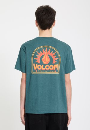 Persoon van achteren gezien die een teal gekleurd shirt met korte mouwen draagt, voorzien van een oranje-geel zonafbeelding en de tekst "VOLCOM True to This" op de achterkant.
