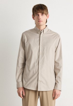 Chemise beige à col avec des fines rayures verticales, manches longues et boutons sur le devant ; présente un petit logo brodé sur la poitrine.