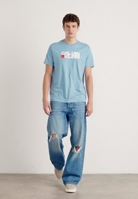 Šviesiai mėlynas medvilninis marškinėlis su balta ir raudona "Calvin Klein" logotipu. Derinamas su nusidėvėjusiais šviesiai mėlynais džinsais ir smėlio spalvos sportbačiais.